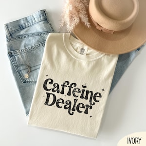May include: An ivory-colored t-shirt with the text "Caffeine Dealer" in a black, distressed font. The shirt is paired with light blue jeans, a beige hat, and a delicate necklace. The shirt has a crew neck and short sleeves.