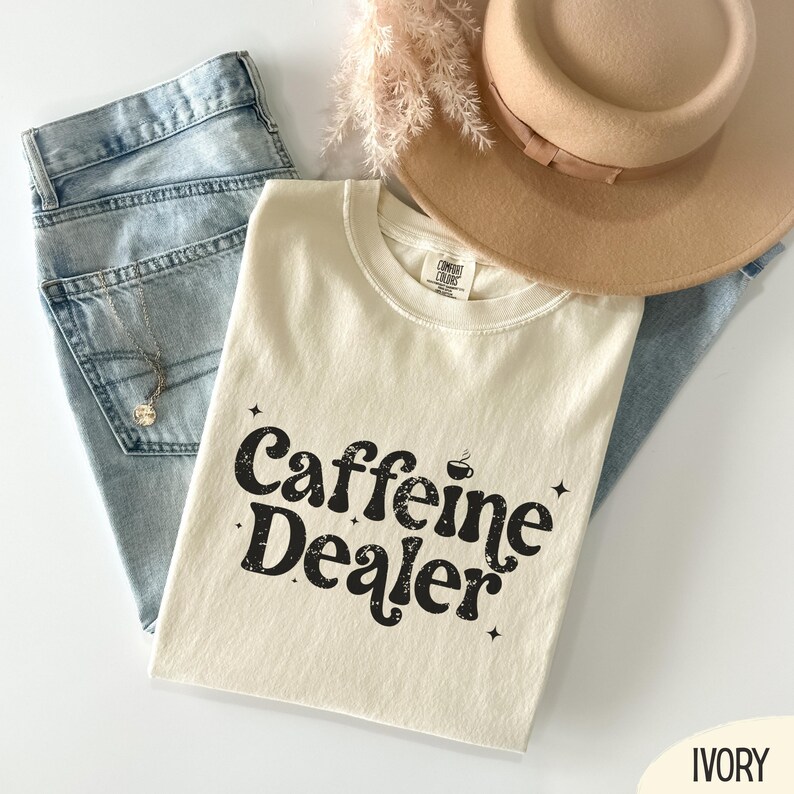 May include: An ivory-colored t-shirt with the text "Caffeine Dealer" in a black, distressed font. The shirt is paired with light blue jeans, a beige hat, and a delicate necklace. The shirt has a crew neck and short sleeves.