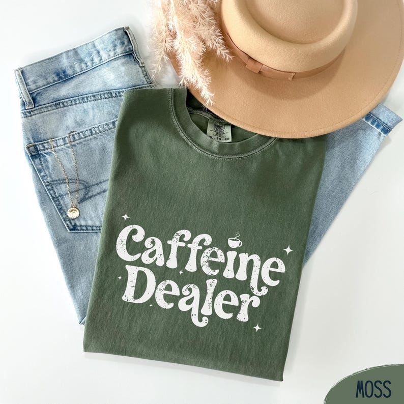 May include: An olive green t-shirt with the text "Caffeine Dealer" in white, along with a coffee cup graphic. The shirt is paired with light blue jeans, a gold necklace, and a tan hat with a brown band.