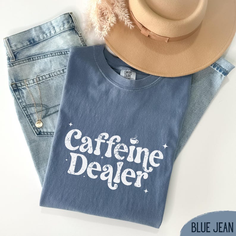 May include: A blue jean Comfort Colors t-shirt with the text "Caffeine Dealer" in white, along with a coffee cup graphic. The shirt is laid on top of a pair of light blue jeans, with a gold necklace and a tan hat.