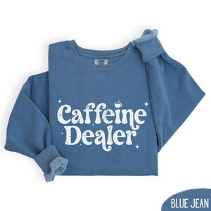 May include: A blue jean colored sweatshirt with the words "Caffeine Dealer" in white, stylized text. The text includes a small coffee cup graphic and starburst accents. The sleeves are folded up.