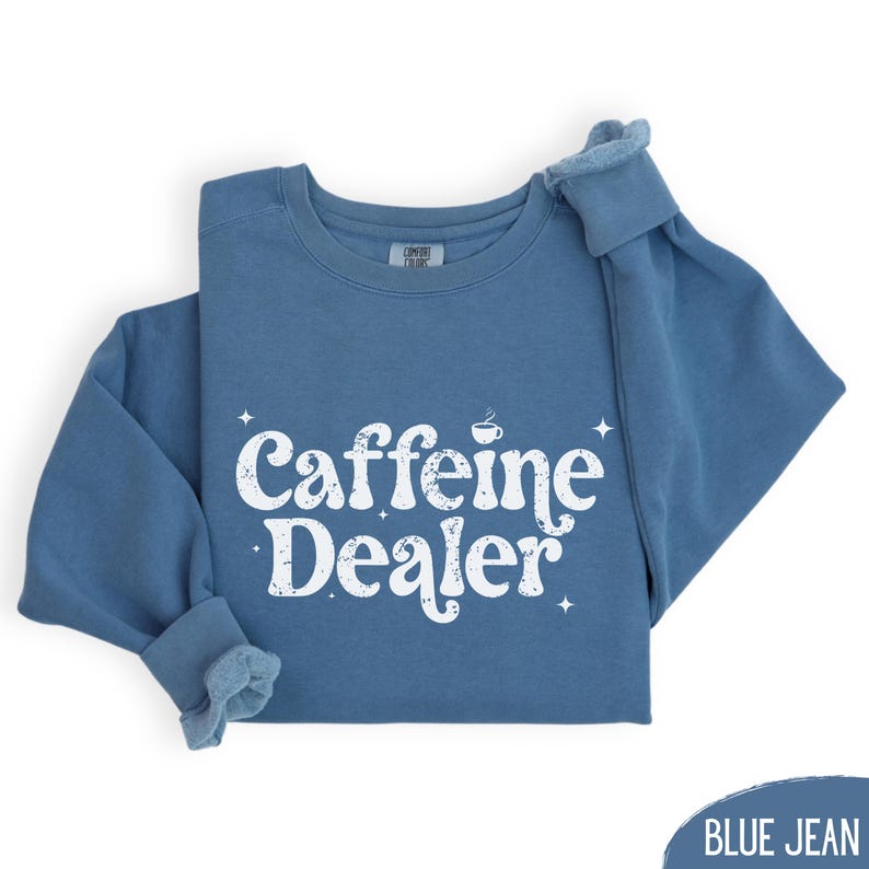 May include: A blue jean colored sweatshirt with the words "Caffeine Dealer" in white, stylized text. The text includes a small coffee cup graphic and starburst accents. The sleeves are folded up.