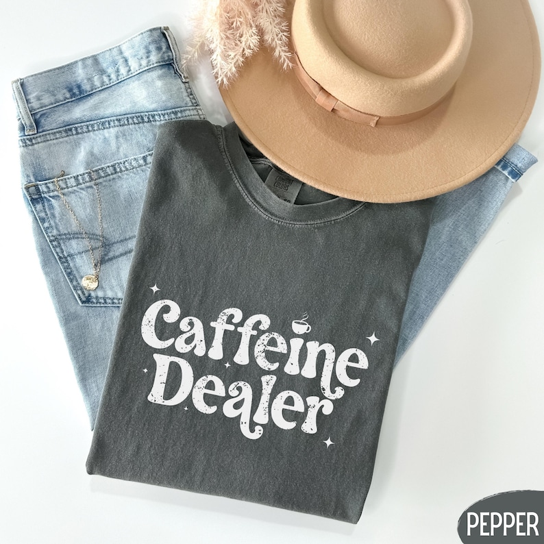 May include: A dark gray t-shirt with the text "Caffeine Dealer" in white, a coffee cup graphic, and star accents. The shirt is laid on top of light blue jeans and a tan hat. A gold necklace is visible.