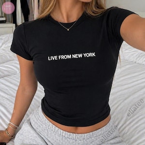 Op de afbeelding: Zwart T-shirt met ronde hals en de witte tekst "LIVE FROM NEW YORK". De top met korte mouwen is gemaakt van een zacht materiaal.