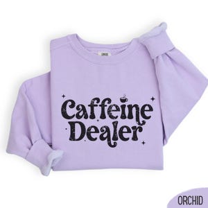 May include: A light purple sweatshirt with the text "Caffeine Dealer" in black. The design includes a coffee cup icon and star accents. The sweatshirt is folded, showing the sleeves and the "COMFORT" label.