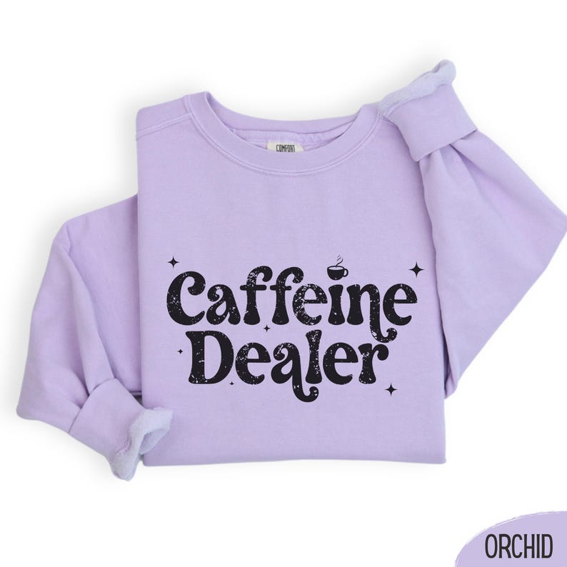 May include: A light purple sweatshirt with the text "Caffeine Dealer" in black. The design includes a coffee cup icon and star accents. The sweatshirt is folded, showing the sleeves and the "COMFORT" label.