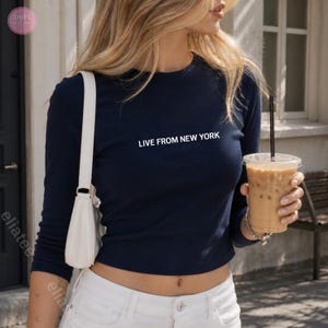 Op de afbeelding: Marineblauw shirt met lange mouwen met de witte tekst "LIVE FROM NEW YORK". Het shirt wordt gecombineerd met een witte broek en een witte schoudertas. De persoon houdt een doorzichtige plastic beker met ijskoffie vast.