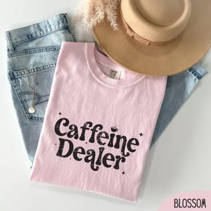 May include: A light pink t-shirt with the text "Caffeine Dealer" in black, along with a small coffee cup graphic. The shirt is laid on top of blue jeans and next to a beige hat.