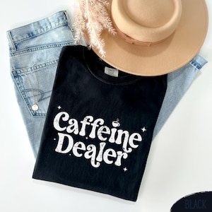 May include: A black t-shirt with the text "Caffeine Dealer" in white, along with a coffee cup graphic. The shirt is laid on top of light blue jeans, a tan hat, and a gold necklace.