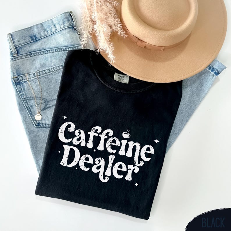 May include: A black t-shirt with the text "Caffeine Dealer" in white, along with a coffee cup graphic. The shirt is laid on top of light blue jeans, a tan hat, and a gold necklace.
