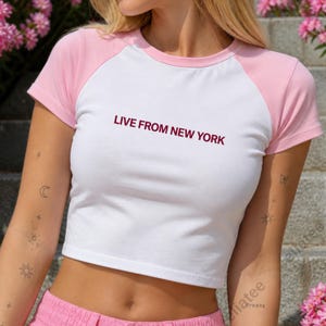 Op de afbeelding: Witte crop top met roze mouwen en de tekst "LIVE FROM NEW YORK" in bordeauxrood. Het shirt heeft een klassiek baseball t-shirt ontwerp, perfect voor casual wear.