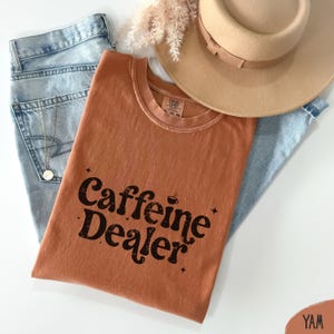 May include: A rust-colored t-shirt with the text "Caffeine Dealer" in a black, retro font. The shirt is folded and rests on top of a pair of light blue jeans and a tan hat. A gold necklace is visible.