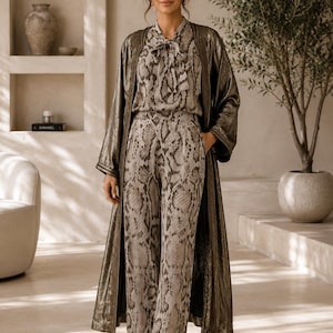Peut inclure: Un ensemble assorti composé d'un chemisier à manches longues avec un col lavallière et d'un pantalon taille haute à jambes larges, tous deux dotés d'un imprimé peau de serpent gris et marron, porté sous un long manteau bronze métallisé.