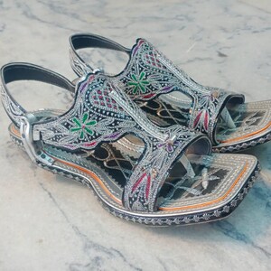 Op de afbeelding: Een paar sierlijke zilveren sandalen met ingewikkelde borduursels. De sandalen hebben een vierkant teenontwerp en zijn versierd met kleurrijke bloemen- en geometrische patronen. De bandjes zijn zwart en de zolen zijn lichtbruin.