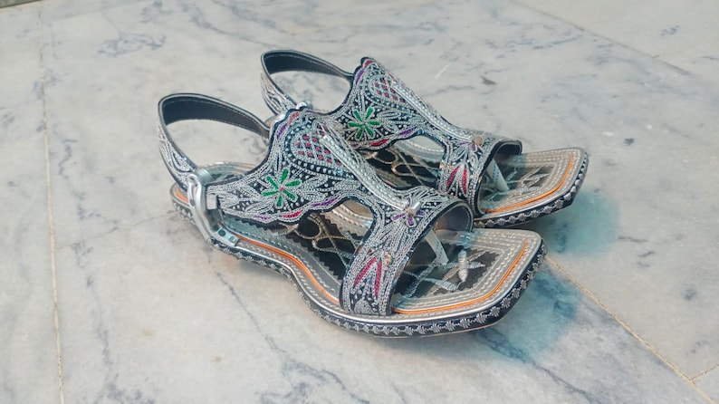 Op de afbeelding: Een paar sierlijke zilveren sandalen met ingewikkelde borduursels. De sandalen hebben een vierkant teenontwerp en zijn versierd met kleurrijke bloemen- en geometrische patronen. De bandjes zijn zwart en de zolen zijn lichtbruin.