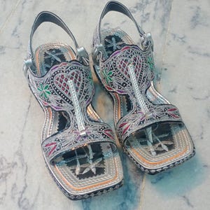 Op de afbeelding: Paar sierlijke zilveren sandalen met ingewikkelde borduursels in rood, groen en paars. De sandalen hebben een T-strap ontwerp en een vierkante neus, met een zilveren band om de enkel. Het schoeisel is waarschijnlijk voor speciale gelegenheden.