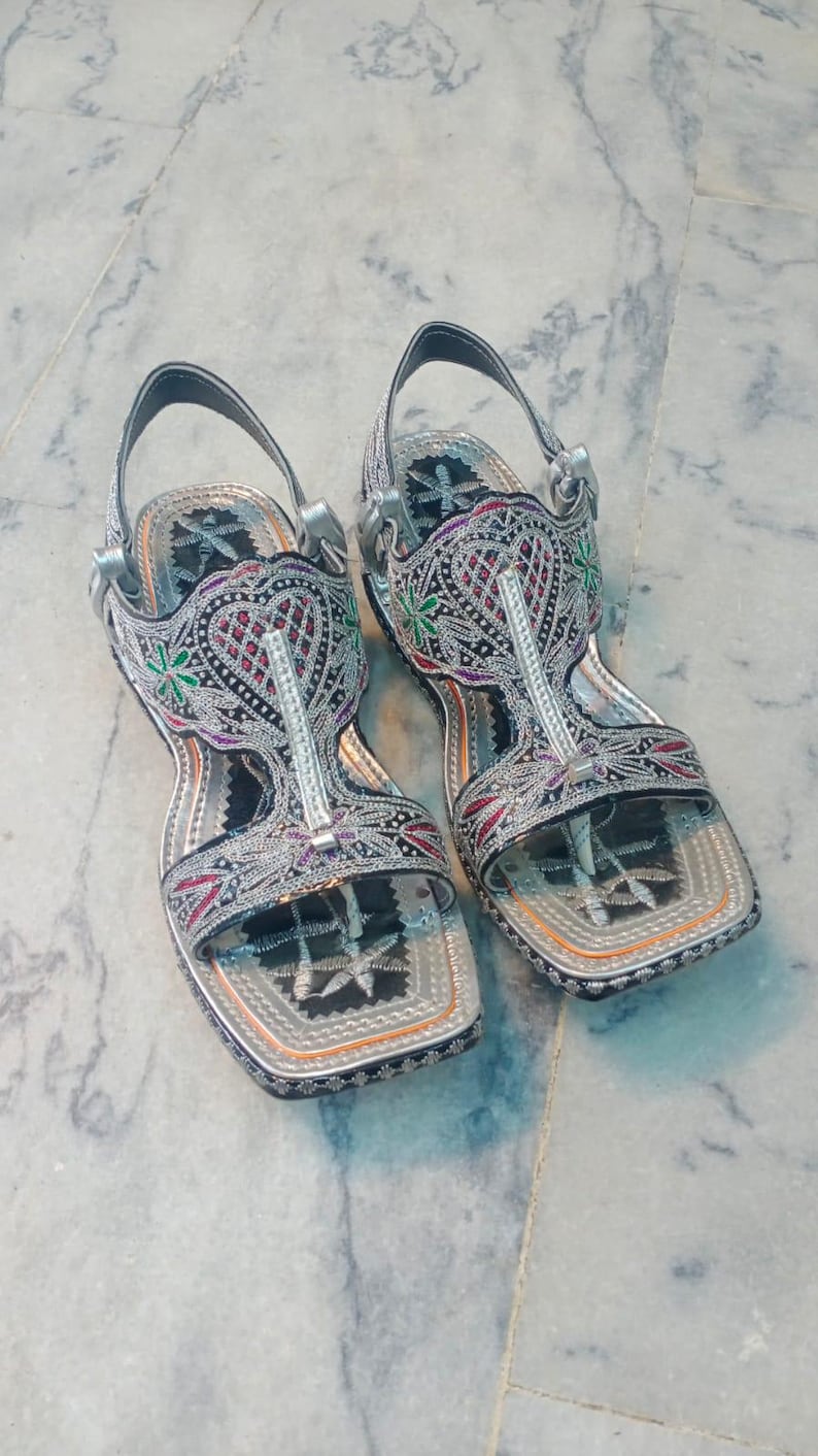 Op de afbeelding: Paar sierlijke zilveren sandalen met ingewikkelde borduursels in rood, groen en paars. De sandalen hebben een T-strap ontwerp en een vierkante neus, met een zilveren band om de enkel. Het schoeisel is waarschijnlijk voor speciale gelegenheden.