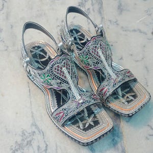 Op de afbeelding: Een paar zilveren sandalen met ingewikkelde borduursels. De sandalen hebben een vierkante neus en een enkelband. De borduursels bevatten rode, groene en zwarte accenten. De sandalen liggen op een marmeren oppervlak.