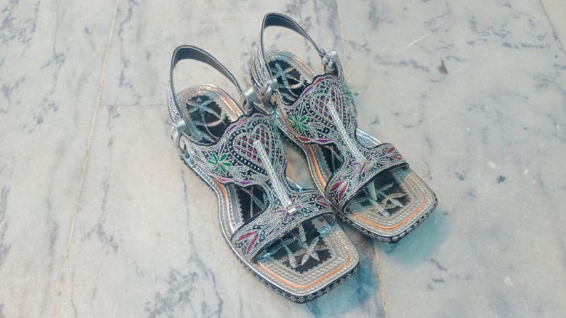Op de afbeelding: Een paar zilveren sandalen met ingewikkelde borduursels. De sandalen hebben een vierkante neus en een enkelband. De borduursels bevatten rode, groene en zwarte accenten. De sandalen liggen op een marmeren oppervlak.