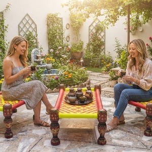 May include: Two rectangular charpai daybeds with bright yellow and red woven rope seats and dark brown carved wooden legs. The furniture is set in a garden courtyard with stone flooring, potted plants, and a small fountain in the background.