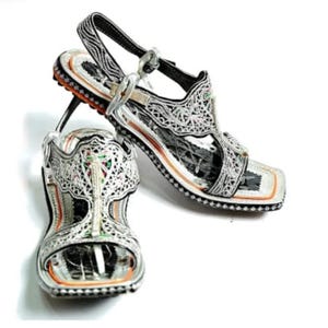 Könnte beinhalten: Ein Paar verzierte silberne Sandalen mit aufwendigen filigranen Designs. Die Sandalen haben eine quadratische Zehenpartie, einen Knöchelriemen und einen dekorativen Absatz. Die Sohlen haben eine schwarz-orangefarbene Umrandung.