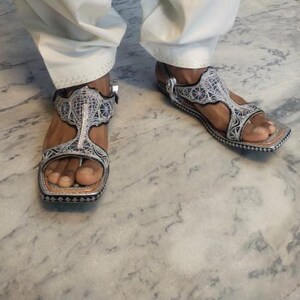 Op de afbeelding: Zilveren en zwarte sandalen met ingewikkelde borduursels en een T-band ontwerp. De sandalen hebben een vierkante neus en een gespsluiting. De zolen zijn donkerbruin. Gedragen met een witte broek.