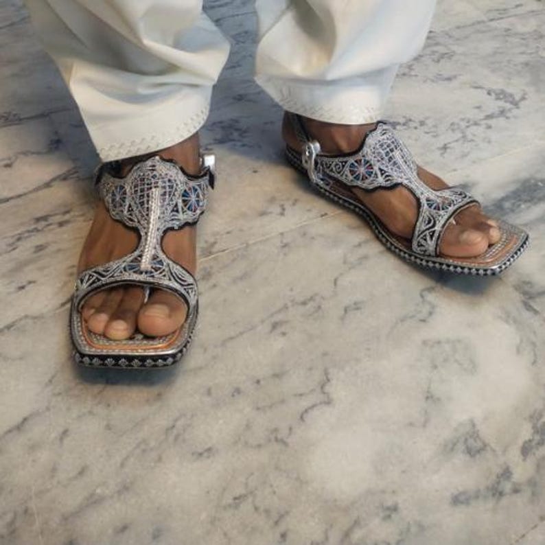 Op de afbeelding: Zilveren en zwarte sandalen met ingewikkelde borduursels en een T-band ontwerp. De sandalen hebben een vierkante neus en een gespsluiting. De zolen zijn donkerbruin. Gedragen met een witte broek.