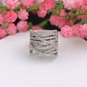 May include: A silver ring featuring a woven design, studded with small, glittering stones. The ring is displayed on a white surface, with pink flowers in the background. The text "HENRY GLORY Handcrafted Jewellery" is visible at the bottom.