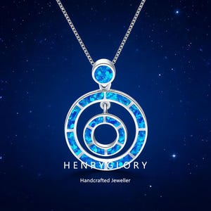 Puede incluir: Un collar de plata con un colgante con un diseño circular con incrustaciones azules tipo ópalo. El colgante tiene tres círculos concéntricos, con un círculo más pequeño suspendido dentro de los más grandes. El texto "HENRY GLORY" y "Handcrafted Jeweller" es visible.