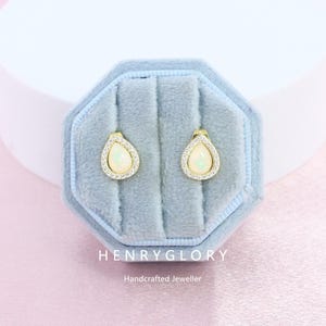 May include: A pair of gold-toned teardrop earrings with a white opal center and a halo of small clear stones. The earrings are displayed in a light blue velvet jewelry box. The box has the text "HENRYGLORY Handcrafted Jeweller".