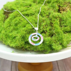 Puede incluir: Un collar de plata con un colgante de doble círculo. El colgante presenta un círculo interior más pequeño dentro de un círculo exterior más grande, ambos adornados con detalles en rosa, azul y blanco. El collar descansa sobre musgo verde.