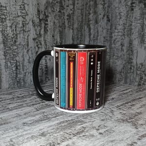 Könnte beinhalten: Eine weiße Keramiktasse mit schwarzem Henkel und Rand. Die Tasse zeigt ein Design von Depeche Mode-Albumcovern, darunter "Some Great Reward" und "Black Celebration". Die Tasse ist ein Artikel zum Thema Musik.