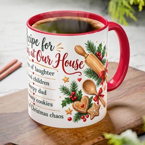 May include: A white ceramic mug with a red interior and handle, featuring the text Recipe for Christmas at Our House, a list of ingredients, and illustrations of a wooden rolling pin, a wooden spoon, gingerbread cookies, and pine branches.