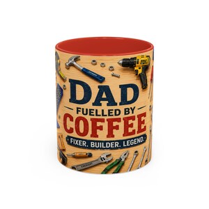 Op de afbeelding: Een keramische mok met een rode binnenkant en een buitenkant met een licht houtnerfpatroon. De mok bevat illustraties van diverse handgereedschappen en elektrisch gereedschap, met de tekst DAD FUELLED BY COFFEE en FIXER. BUILDER. LEGEND. in blauw en rood.