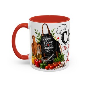 Può includere: Una tazza in ceramica bianca con manico e bordo interno rossi. L'esterno presenta un grembiule nero con il testo "GOOD FOOD GOOD MOOD", un cappello da chef, un tagliere, olio, peperoni, aglio e la scritta "CHEF The Secret Is Love."