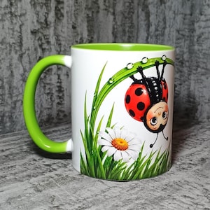 Op de afbeelding: Een witte keramische mok met een felgroen handvat en binnenkant. De buitenkant is voorzien van een grafische illustratie van een rood lieveheersbeestje met zwarte stippen dat ondersteboven aan een groen grassprietje met waterdruppels hangt.