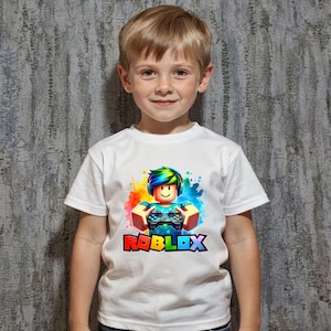 May include: White t-shirt featuring a colorful graphic of a Roblox character holding a game controller, with the Roblox logo in rainbow colors. The shirt is worn by a young child.