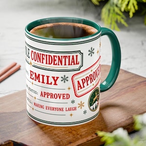 Op de afbeelding: Een witte keramische mok met een donkergroene binnenkant en handvat, voorzien van rode en groene tekst "ROLE CONFIDENTIAL", "EMILY", "APPROVED" en "SPECIAL SKILL: MAKING EVERYONE LAUGH", geaccentueerd met sneeuwvlok- en stergraphics.