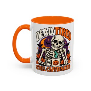 Könnte beinhalten: Ein weißer Keramikbecher mit orangefarbenem Henkel und Innenraum, verziert mit einem Skelett, das den Kopf auf eine Hand stützt und eine kleine Kürbistasse hält. Der Text DEAD TIRED STILL CAFFEINATED ist von Fledermäusen und Herbstblättern umgeben.