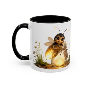 Op de afbeelding: Een witte keramische mok met een zwarte handgreep en binnenrand. De buitenkant toont een afbeelding van een cartoonbij die op een glazen pot vol gloeiend licht zit. De pot is vastgebonden met touw en een klein bruin label met een getekend hart.
