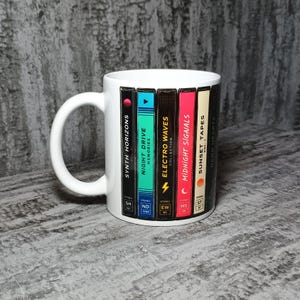 May include: White ceramic mug featuring a design of colorful cassette tapes. The tapes are labeled "Synth Horizons," "Night Drive Memories," "Electro Waves Collection," "Midnight Signals," and "Sunset Tapes." The mug has a white handle.