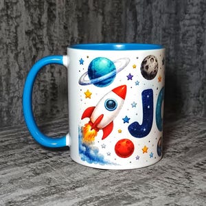 Pode incluir: Uma caneca de cerâmica branca com uma alça e borda interna azul brilhante. O exterior apresenta uma ilustração colorida com tema espacial, incluindo um foguete, planetas, estrelas e a letra J azul parcial. O design inclui um foguete vermelho.