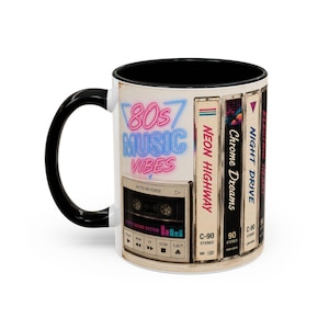 Op de afbeelding: Witte keramische mok met een zwarte handgreep en binnenkant. De mok heeft een ontwerp met de tekst "80s Music Vibes" in neonkleuren, samen met illustraties van cassettes en de tekst "Neon Highway", "Chrome Dreams" en "Night Drive".