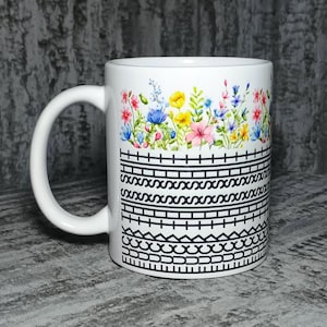 Puede incluir: Una taza de cerámica blanca con un diseño floral de acuarela colorido en la parte superior, que muestra varias flores pequeñas en tonos rosa, amarillo y azul. Debajo de las flores, la taza presenta una serie de patrones y líneas geométricas negras.