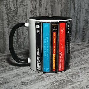 Op de afbeelding: Een witte keramische mok met een zwarte handgreep en binnenkant, voorzien van een grafische print van vijf verticaal geplaatste Depeche Mode-cassettes. De labels tonen de albumtitels Speak & Spell, Some Great Reward, Black Celebration, Music for the Masses en Violator.