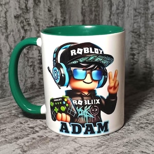 Peut inclure: Un mug en céramique blanche avec une anse et un intérieur vert foncé. L'avant présente un personnage Roblox portant une casquette noire, des lunettes de soleil bleues et un casque, tenant une manette. Le nom ADAM est imprimé en bleu en dessous.