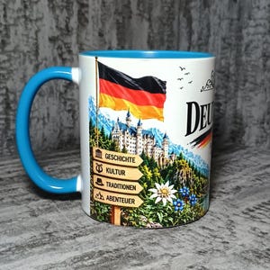Puede incluir: Una taza de cerámica blanca con asa e interior de color azul brillante. El exterior presenta una bandera alemana, un castillo y una señal de madera con las palabras Geschichte, Kultur, Traditionen y Abenteuer, junto a edelweiss y flores azules.