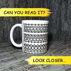Może przedstawiać: Biały ceramiczny kubek z czarnym wzorem graficznym składającym się z poziomych rzędów geometrycznych kształtów, w tym krzyżyków, prostokątów i pętli. Napis CAN YOU READ IT? znajduje się na górze, a LOOK CLOSER... na dole czcionką.