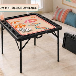 May include: A black folding game table with a beige mat featuring a beach-themed print of umbrellas and lounge chairs. The table has built-in cup holders and storage slots. A text box reads CUSTOM MAT DESIGN AVAILABLE. A folded table stands nearby.