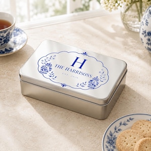 May include: A rectangular silver metal tin with a hinged lid featuring a blue printed design. The design includes a central letter H, the text THE HARRISONS, and EST. 2026, surrounded by a decorative border with blue floral ginger jar motifs.